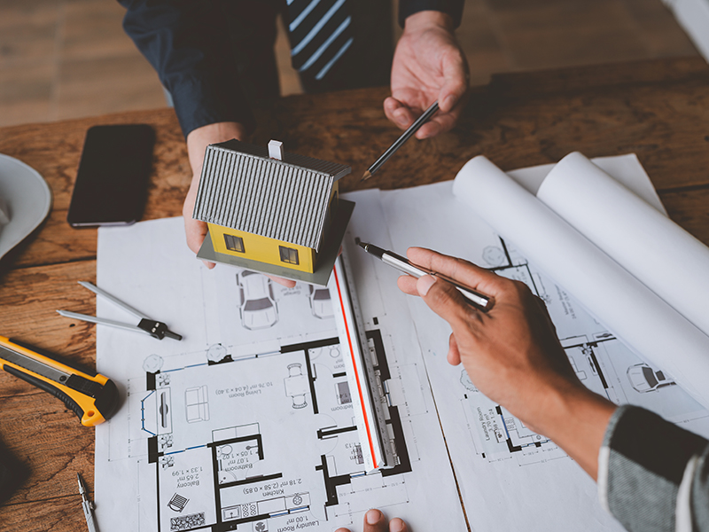 Personen diskutieren über Baupläne, während ein Modellhaus auf dem Tisch steht, umgeben von Werkzeugen und Zeichnungen.