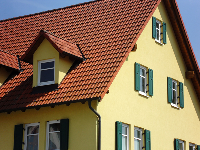 Dachansicht eines gelben Hauses mit roten Ziegeln und grünen Fensterläden unter blauem Himmel.