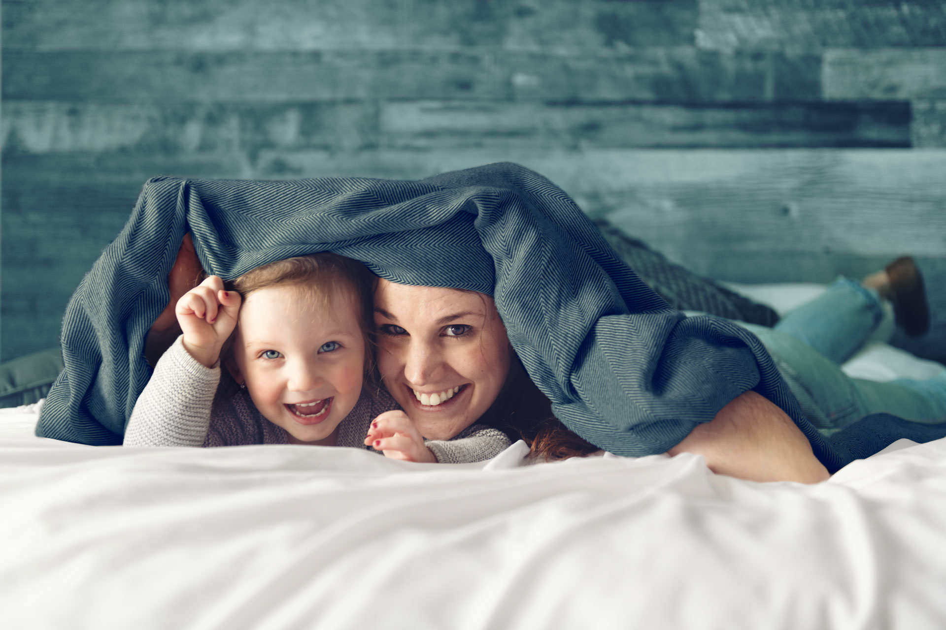Mutter und Tochter lächeln unter einer Decke auf dem Bett, umgeben von einer gemütlichen Atmosphäre.