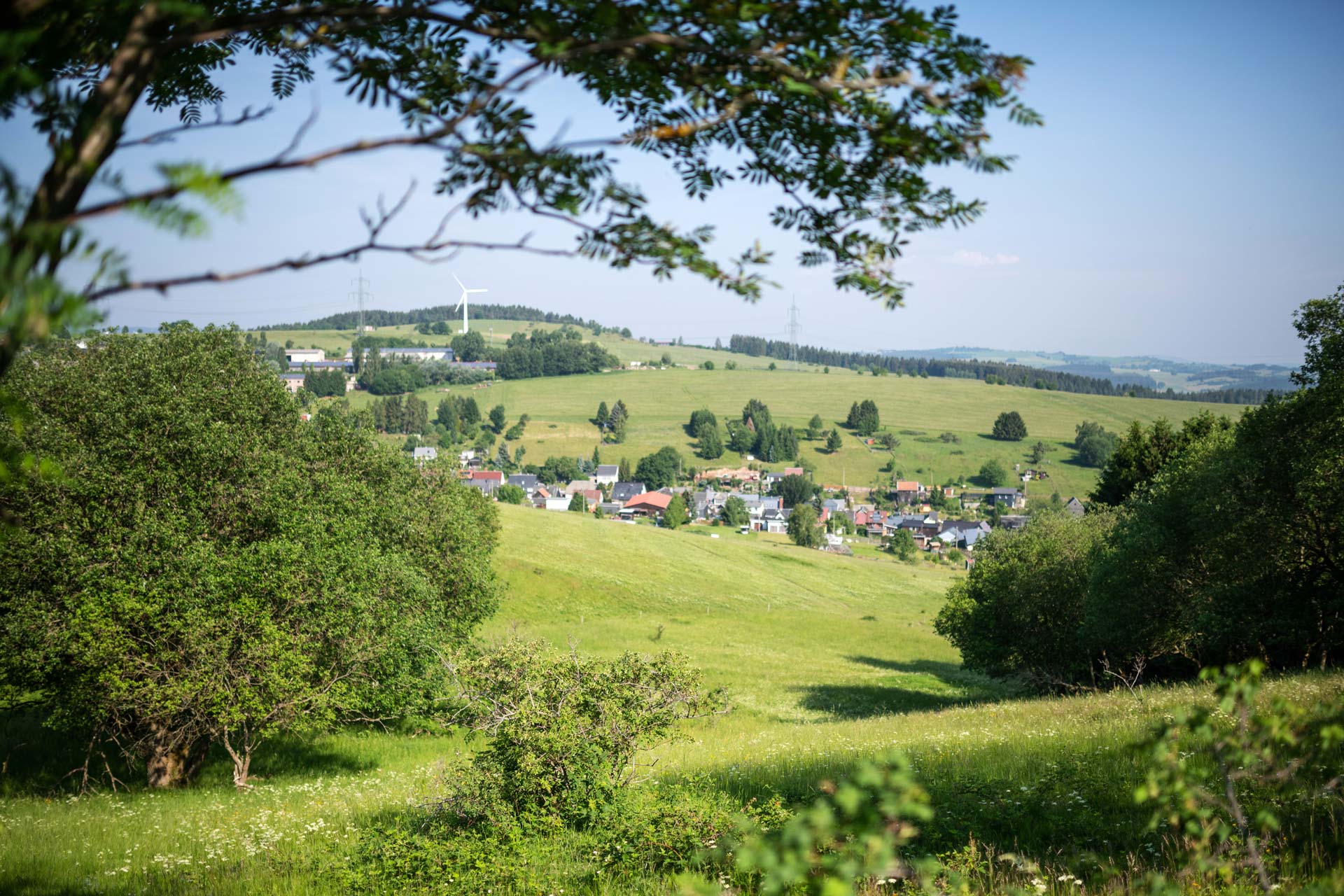Landschaft mit weiten Wiesen, Bäumen und einem Dorf im Hintergrund, umgeben von sanften Hügeln und Windrädern.
