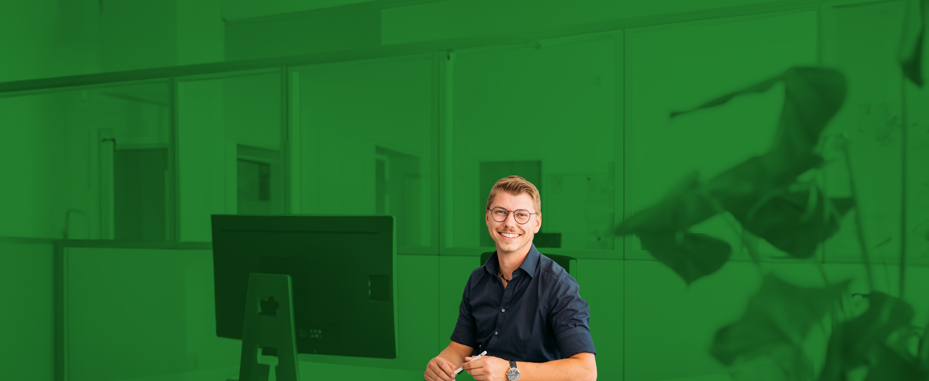 Lächelnder Mann mit Brille sitzt an einem Schreibtisch in einem modernen Büro, umgeben von Pflanzen.