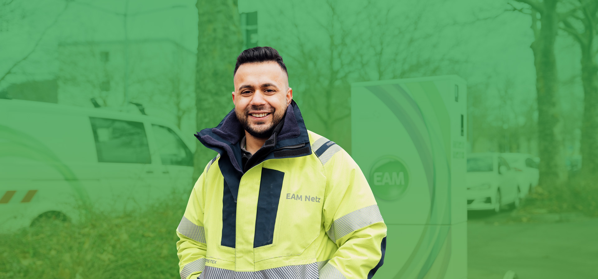 Ein lächelnder Mann in gelber Arbeitsbekleidung steht im Freien, umgeben von Bäumen und einem Fahrzeug.