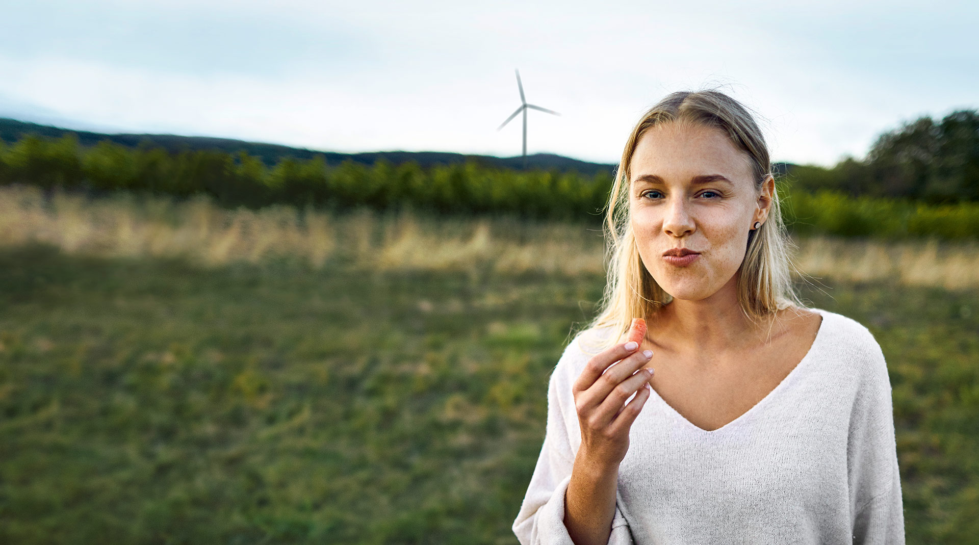 Frau mit blonden Haaren, die in einem Feld steht und genüsslich einen Snack isst, Windrad im Hintergrund.