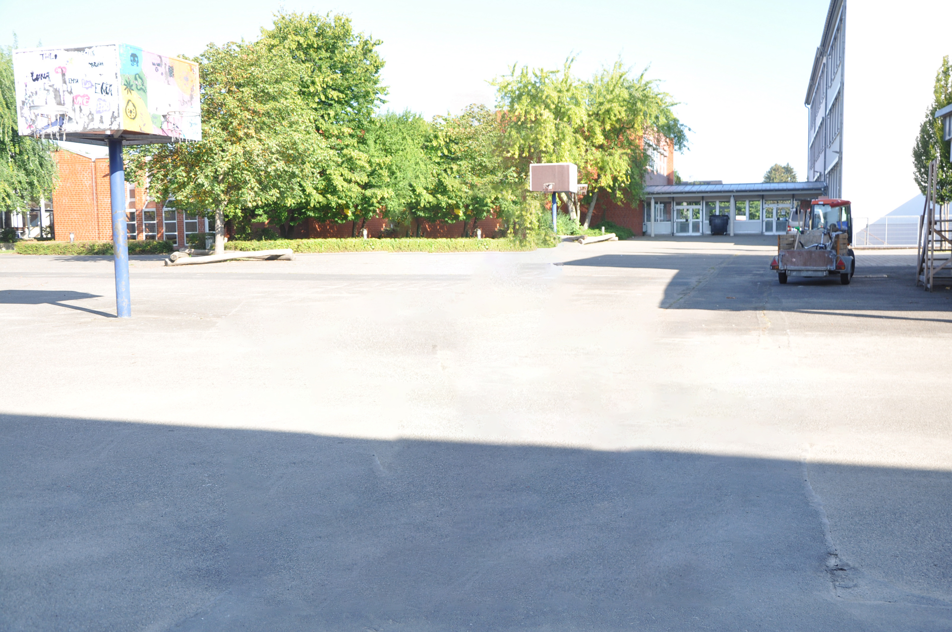 Weitläufiger Schulhof mit Basketballkorb, umgeben von Bäumen und einer Schulgebäude im Hintergrund. Ein Fahrzeug steht auf der Fläche.