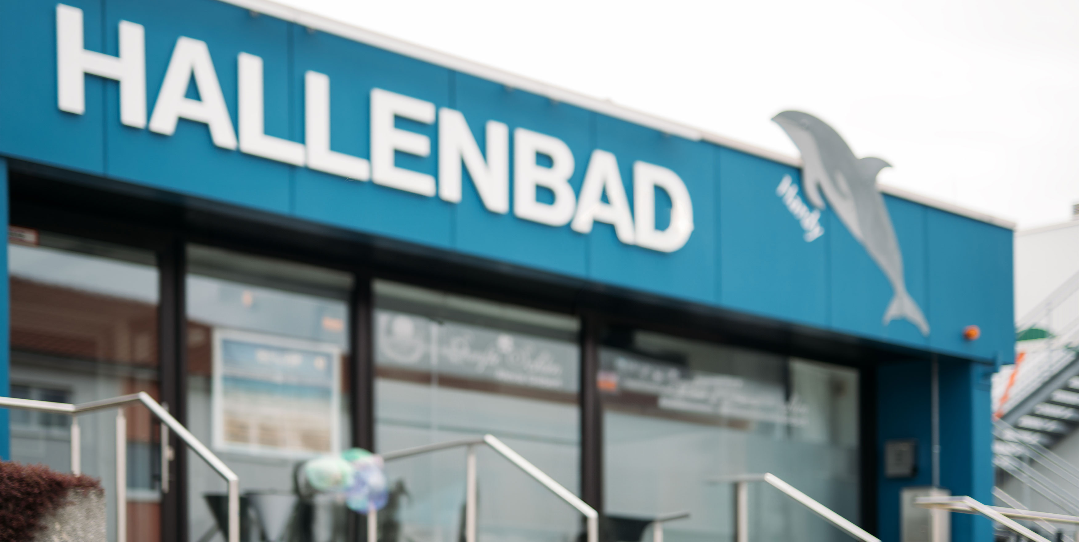 Eingang zum Hallenbad mit blauer Fassade und weißen Buchstaben, im Hintergrund eine Treppe und ein Delphin-Symbol.