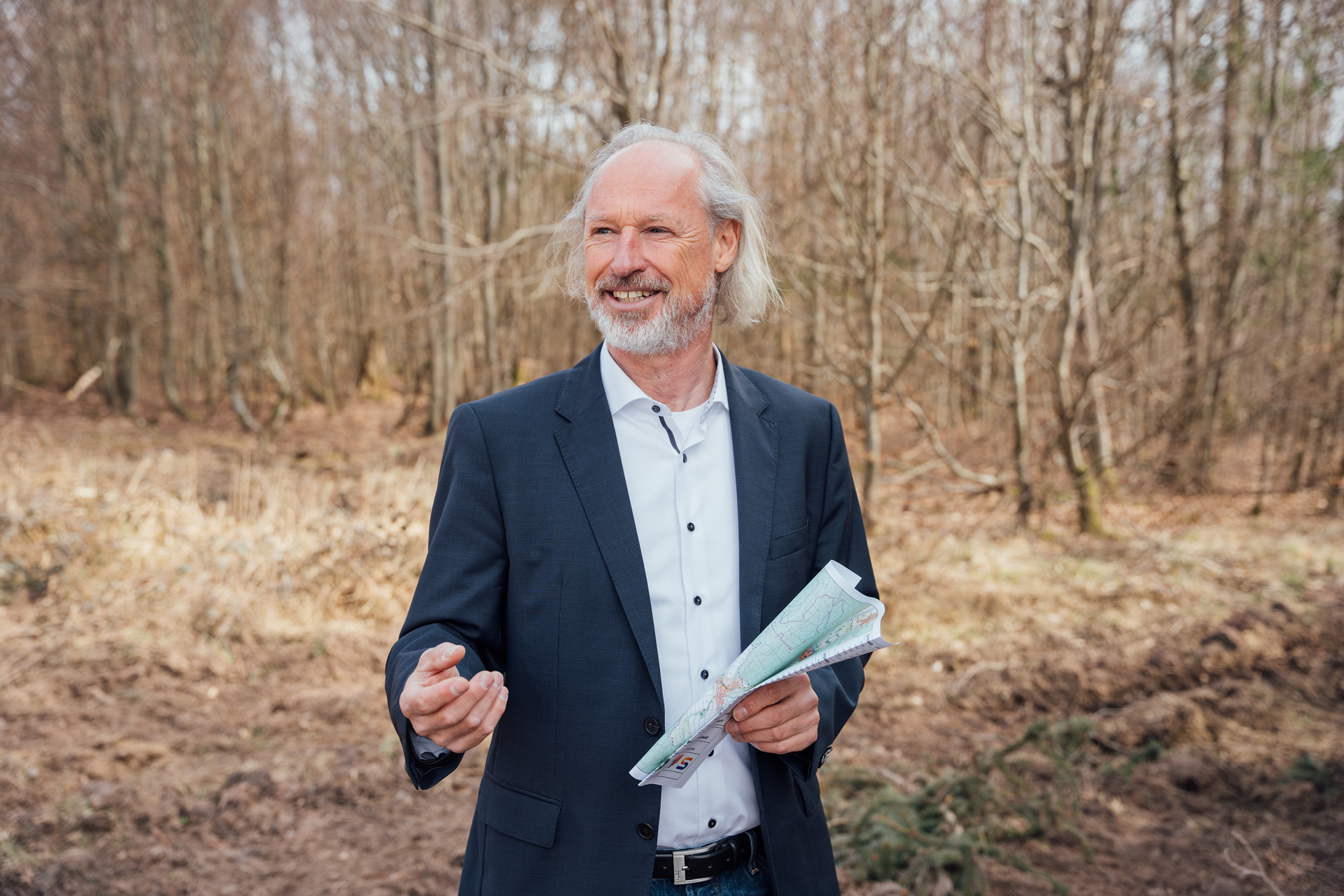Mann im Anzug im Wald, der eine Karte in der Hand hält und mit gestikulierender Hand spricht.