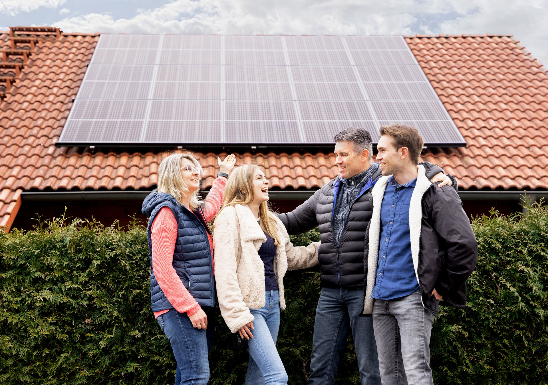 Familie steht vor einem Haus mit Solarzellen auf dem Dach und lacht fröhlich miteinander.
