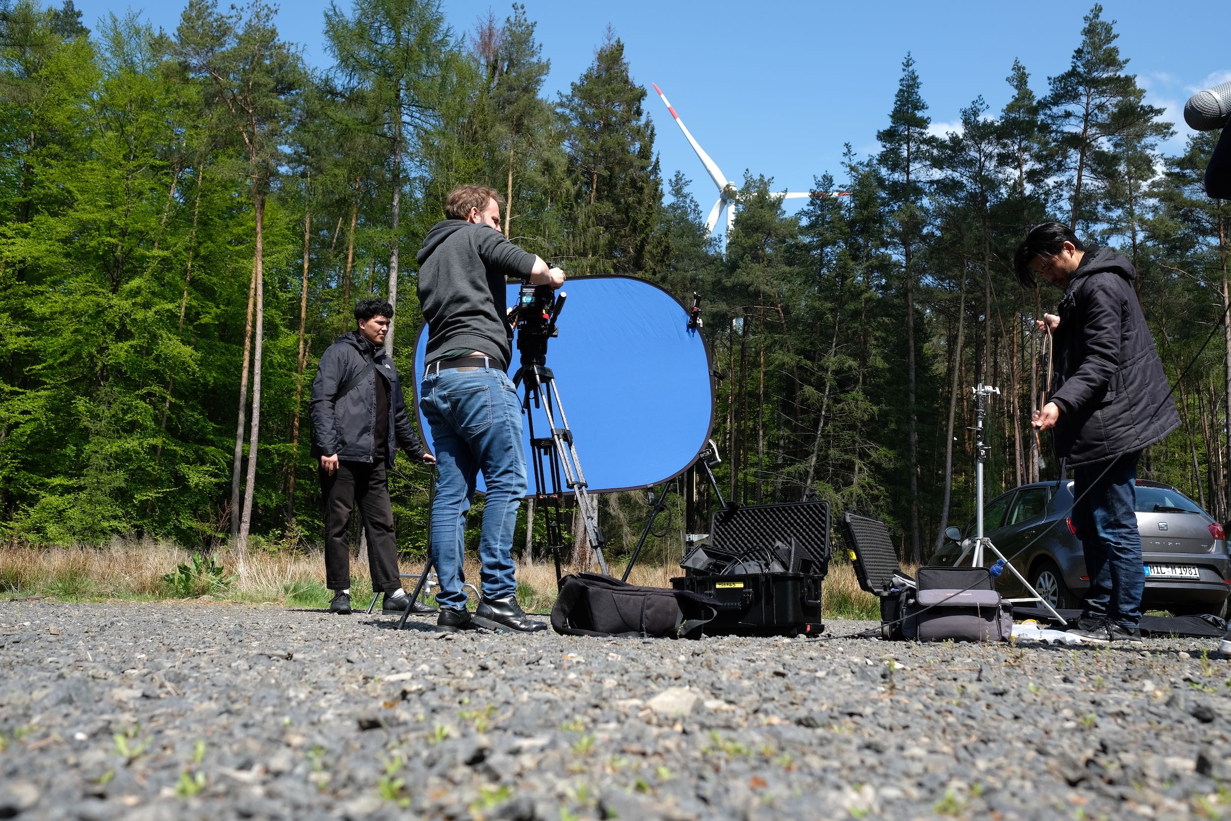 Zwei Personen bearbeiten eine Filmszene im Freien, umgeben von Bäumen, mit Kameratechnik und einem blauen Hintergrund.