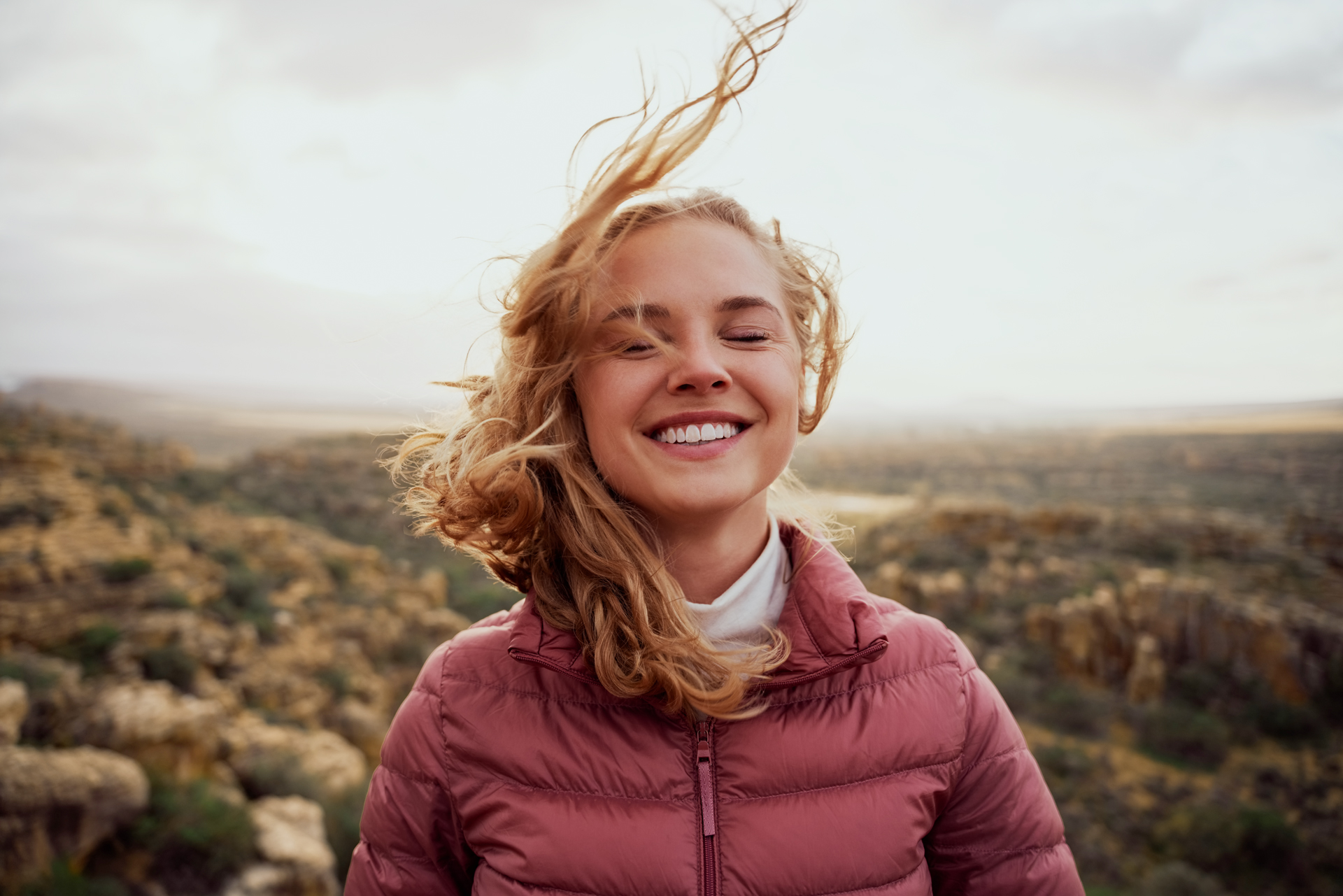 Lächelnde Frau mit lockigem Haar, die in einer natürlichen Landschaft steht, mit sanften Winden, die ihre Haare bewegen.