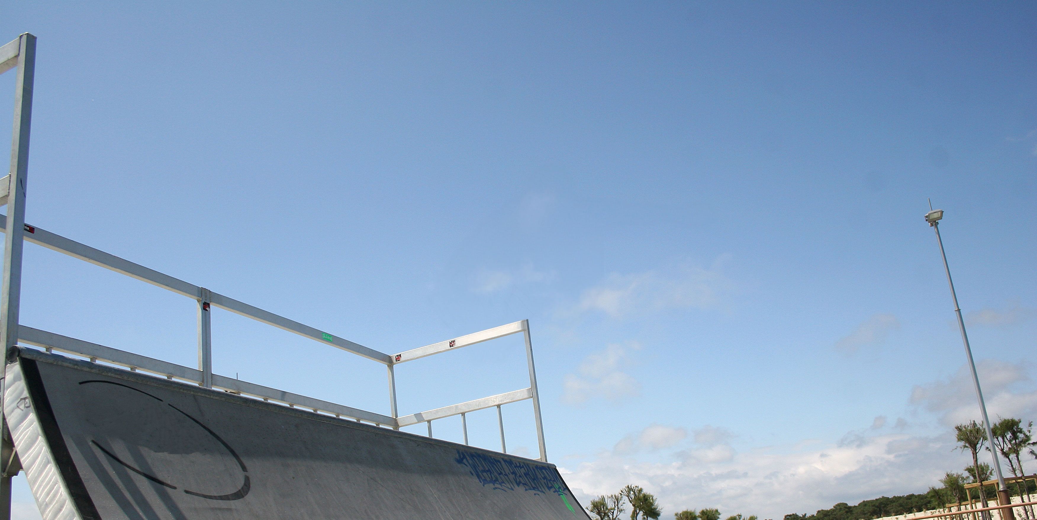 Ein Skatepark-Rampe unter blue ciel, mit Metallgeländer und einer Straßenlaterne im Hintergrund.