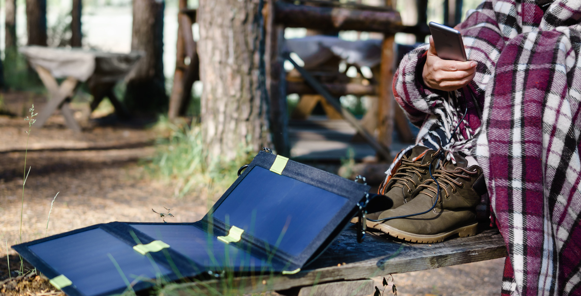 Person in Decke sitzt auf einer Bank, nutzt Smartphone, neben ihnen liegen Wanderschuhe und ein aufgerolltes Solarpanel.