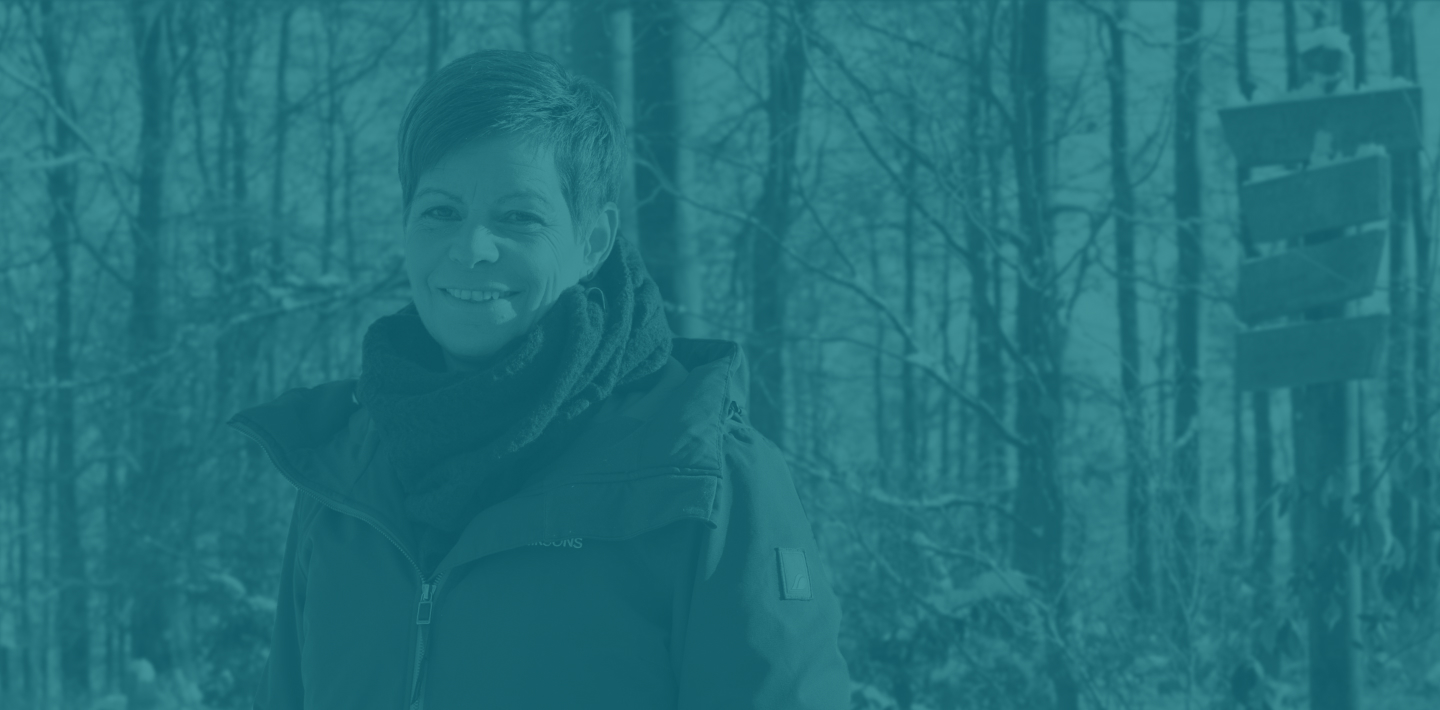 Frau mit kurzer Frisur in warmer Jacke und Schal, lächelt im verschneiten Wald hinter einem Schild.