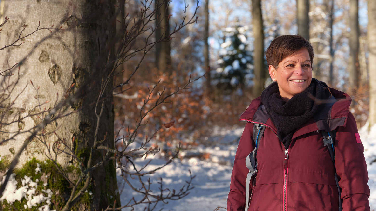 Frau lächelt im Winterwald, umgeben von schneebedeckten Bäumen und einer warmen Jacke, vor einem großen Baumstamm.