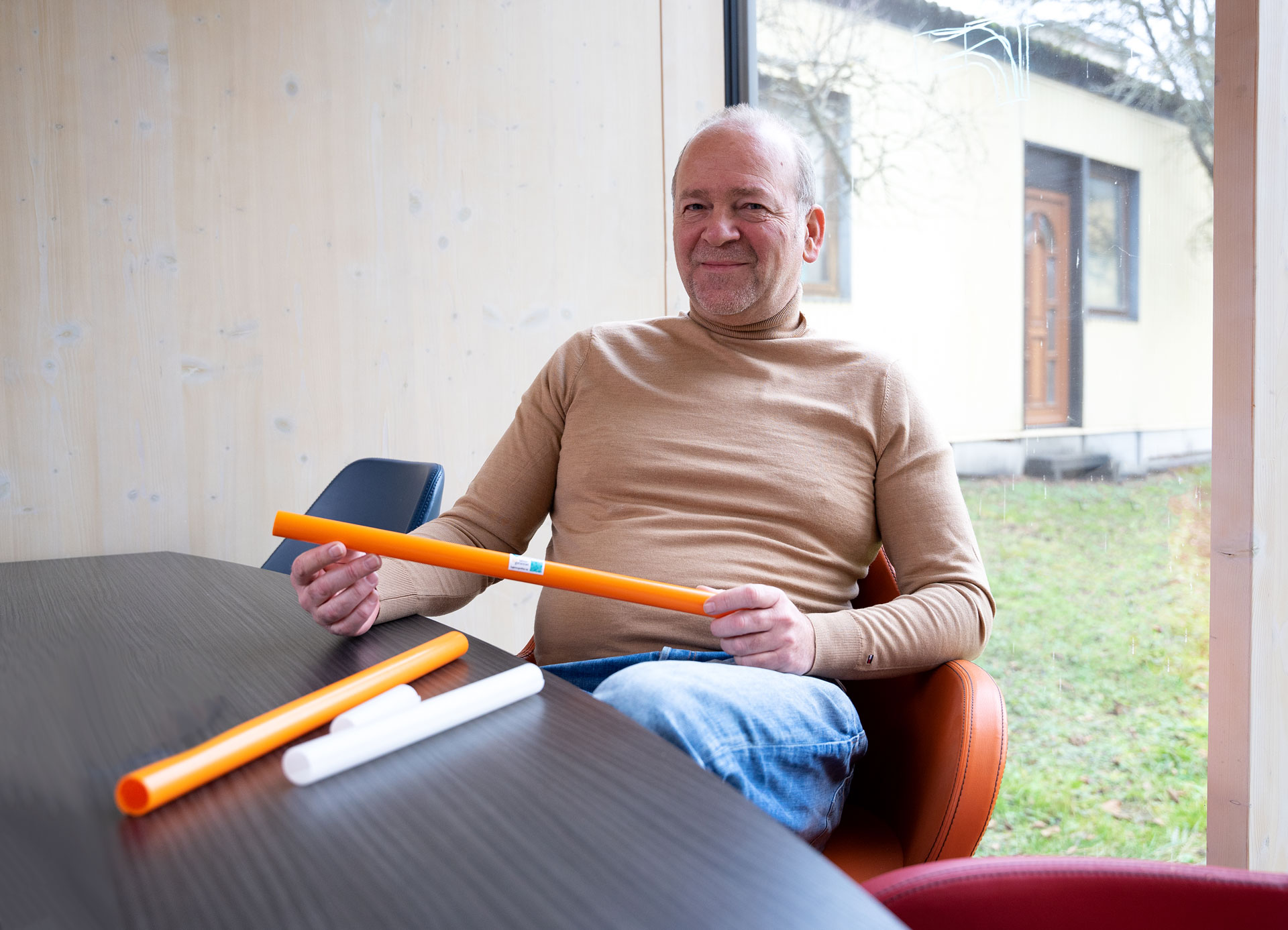 Mann im Pulli hält orangefarbene Rohre, sitzt an einem Tisch mit weißen Rohren, Blick aus einem Fenster auf einen Garten.
