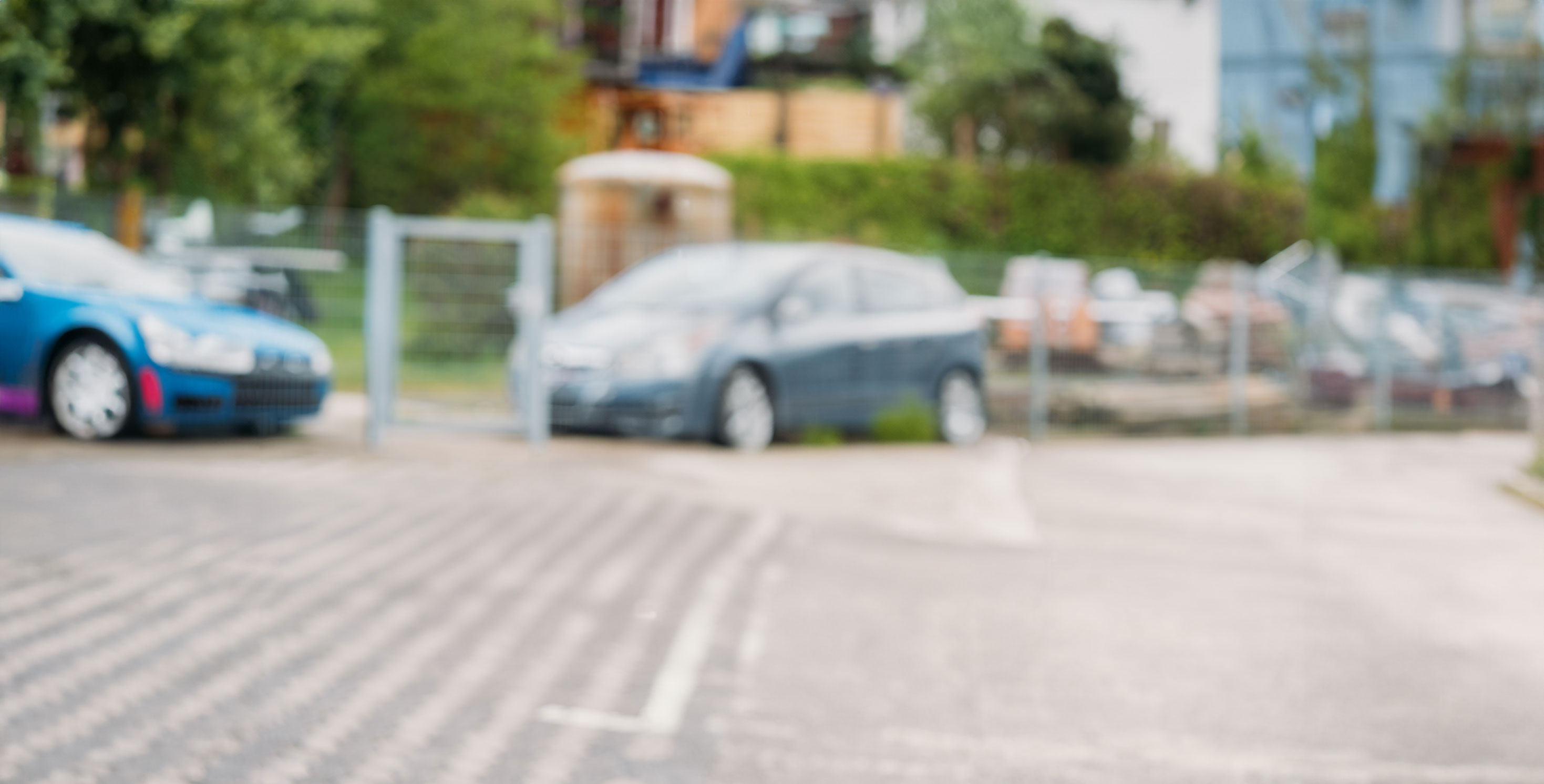 Unscharfes Bild eines Parkplatzes mit zwei parkenden Autos und einer umgebenden Zaunanlage.