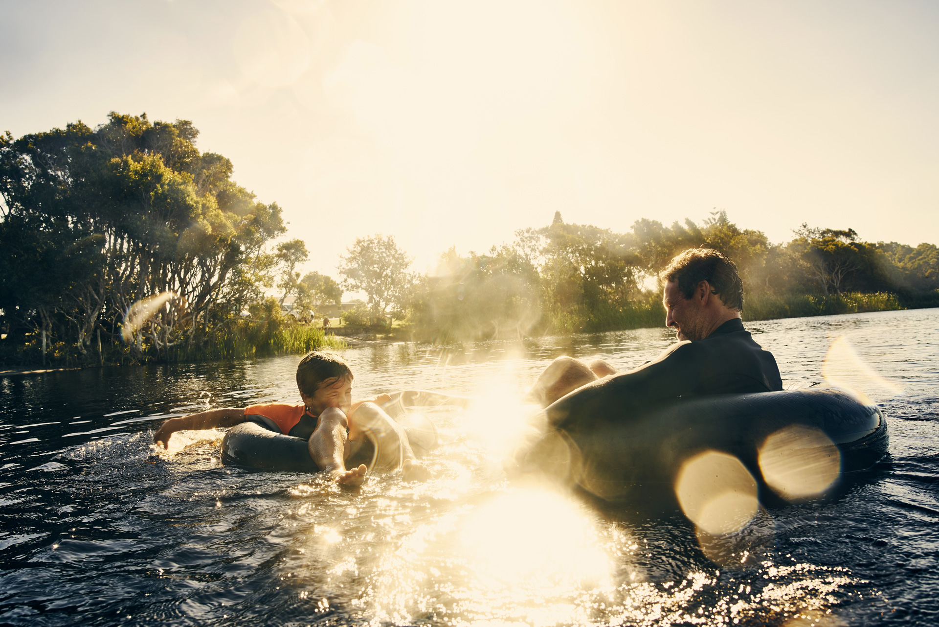Zwei Personen im Wasser, die auf Gummireifen entspannen, umgeben von natürlicher Landschaft und Sonnenlicht.