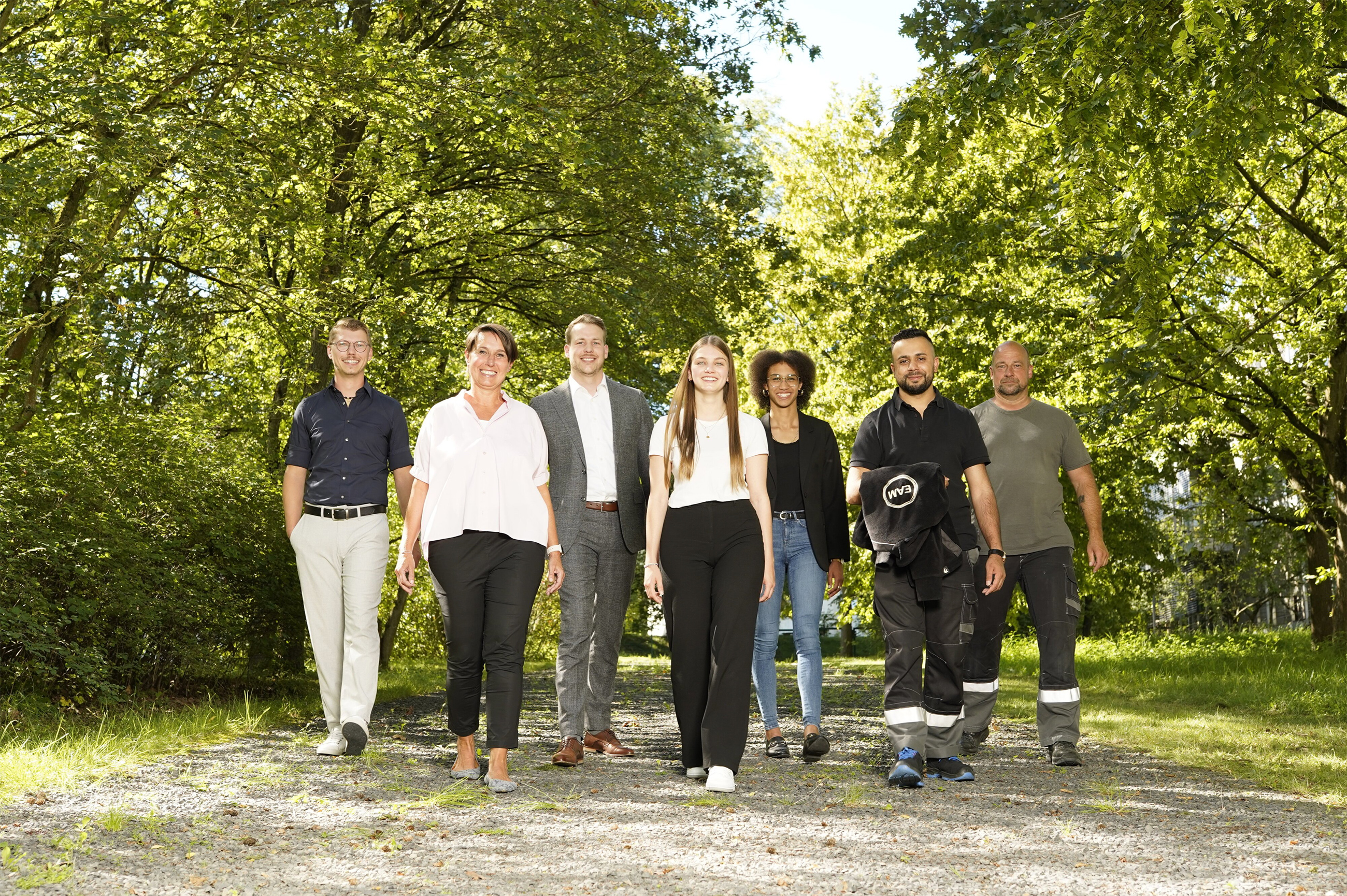 Sechs Personen in Arbeitskleidung und Freizeitkleidung laufen auf einem Kiesweg durch einen bepflanzten Park.
