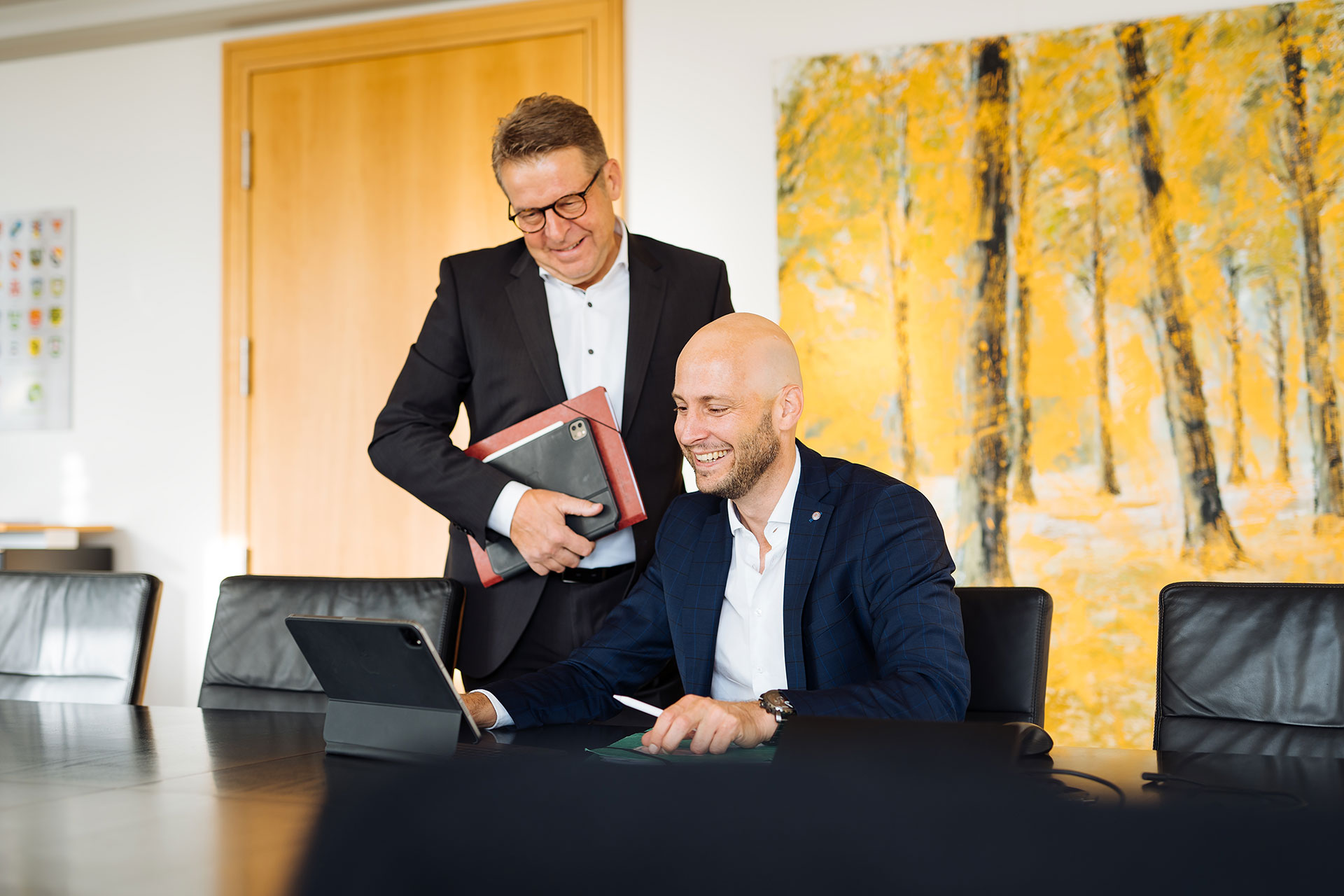 Zwei Personen in einem modern gestalteten Besprechungsraum, eine Person arbeitet an einem Tablet, die andere betrachtet sie lächelnd.