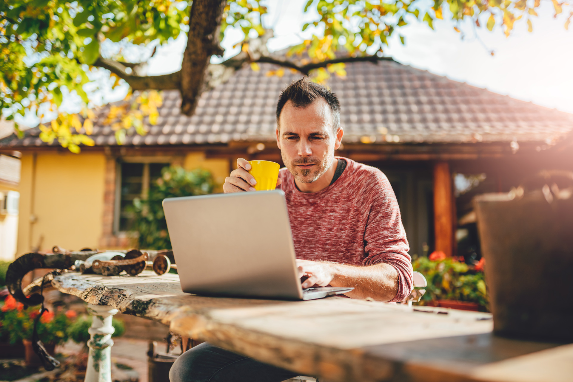 Mann sitzt an einem Tisch im Freien, arbeitet am Laptop und hält eine gelbe Tasse in der Hand.